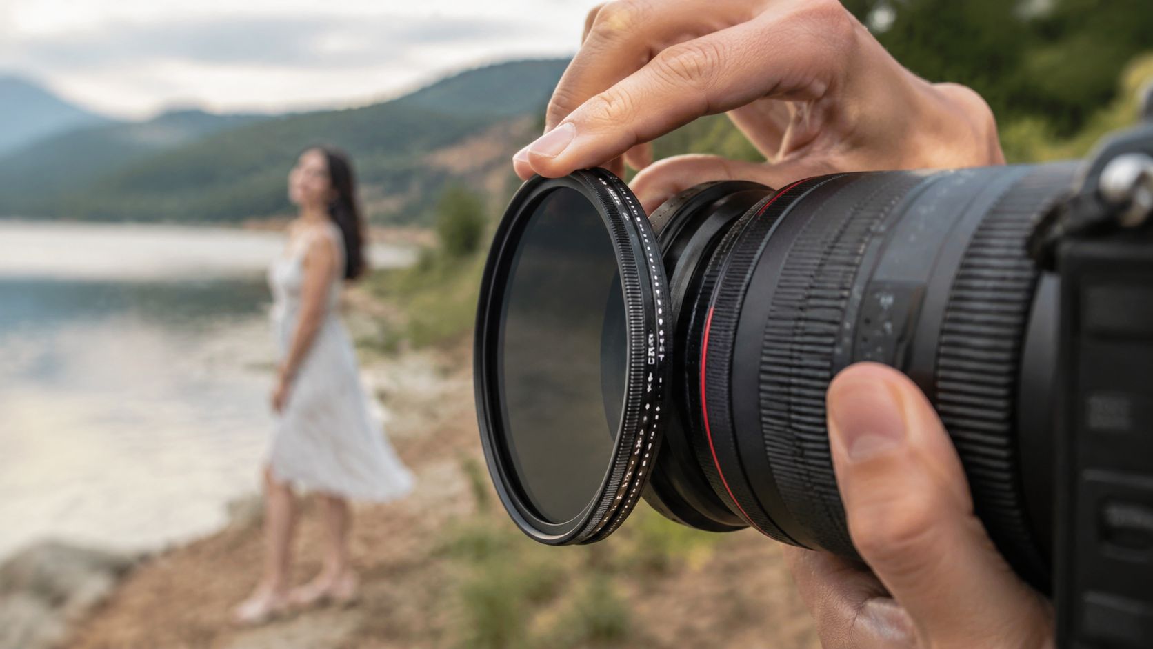 A photographer's hand attaching a circular polarizing filter to a camera lens outdoors near a lake.