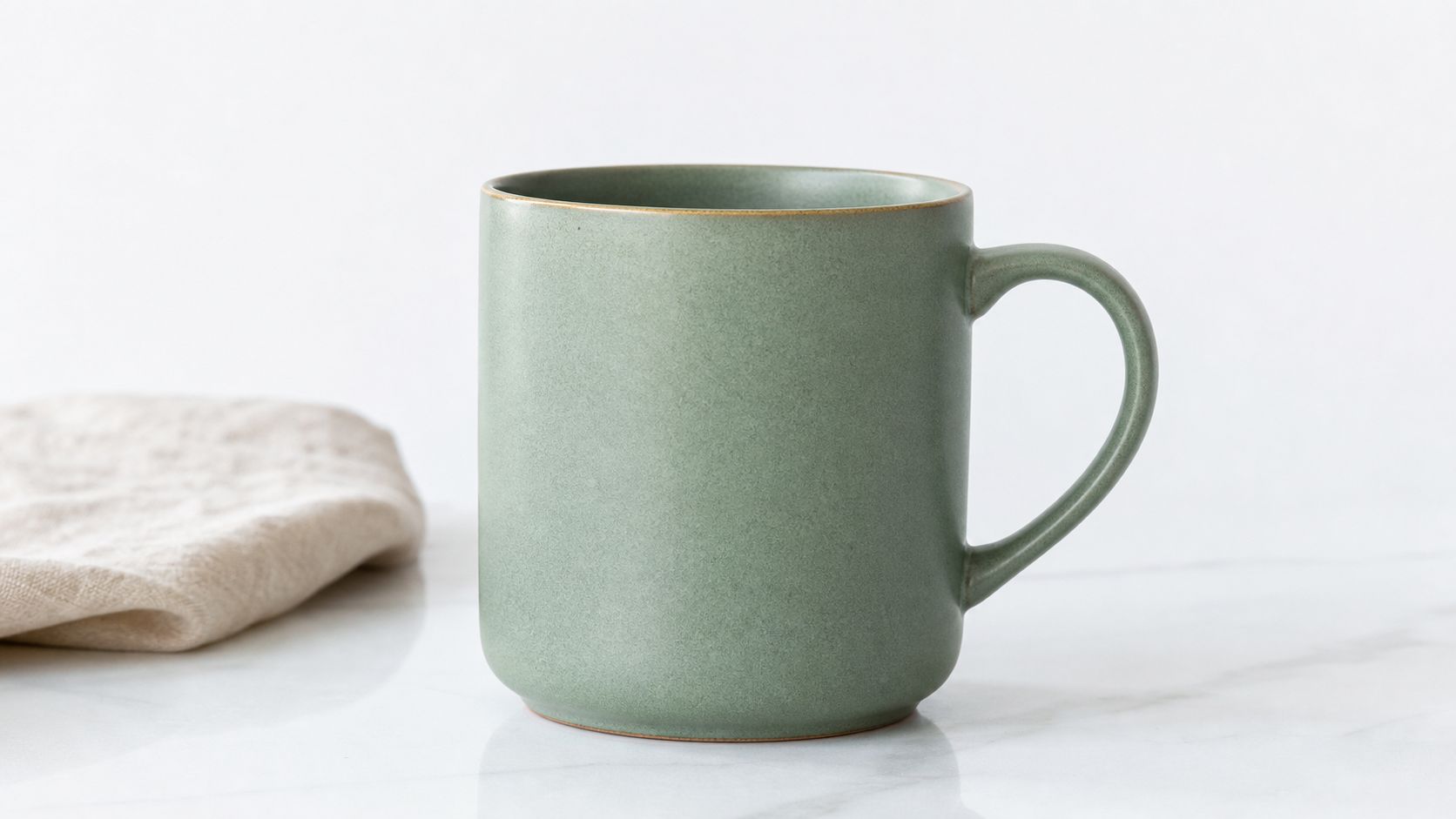 A sage green ceramic coffee mug sitting on a white marble surface next to a beige cloth.